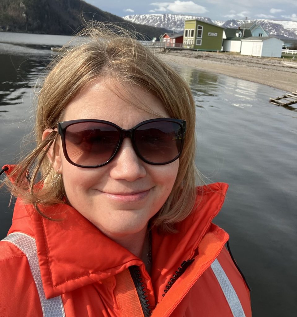Photo of white woman with light brown hair smiling and wearing sunglasses and a bright orange jacket